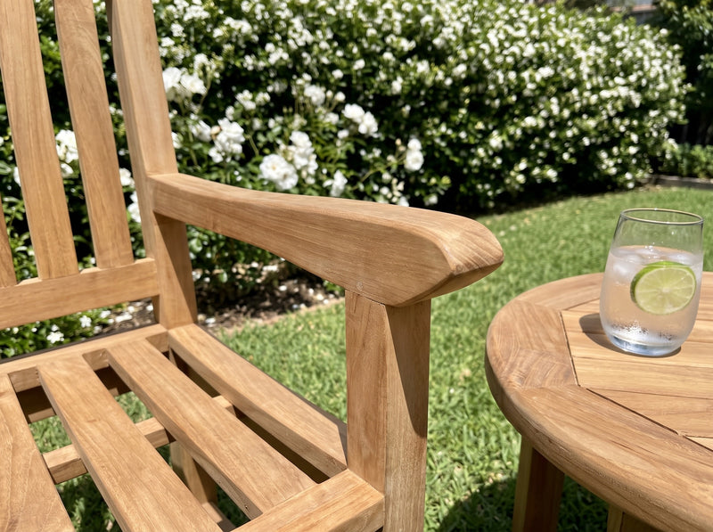 Close-up of the broad armrest of a teak Sierra armchair beside a round teak side table in a sunny garden