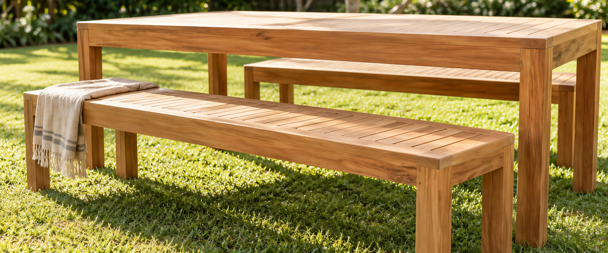 Two teak dining benches at a teak table on a sunlit lawn
