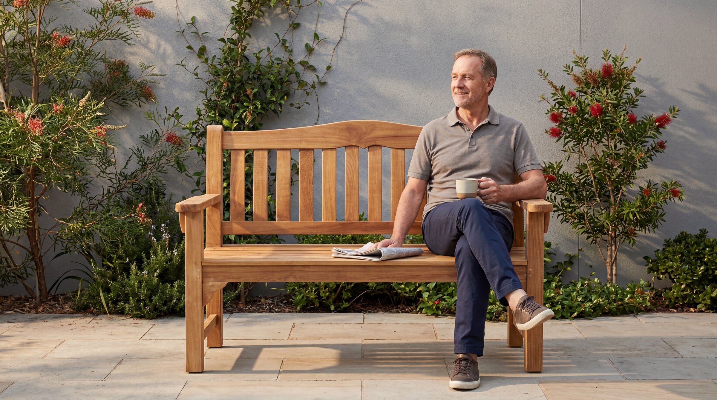 A relaxed man enjoying coffee and a newspaper on a Glasgow teak garden bench in a landscaped outdoor setting.