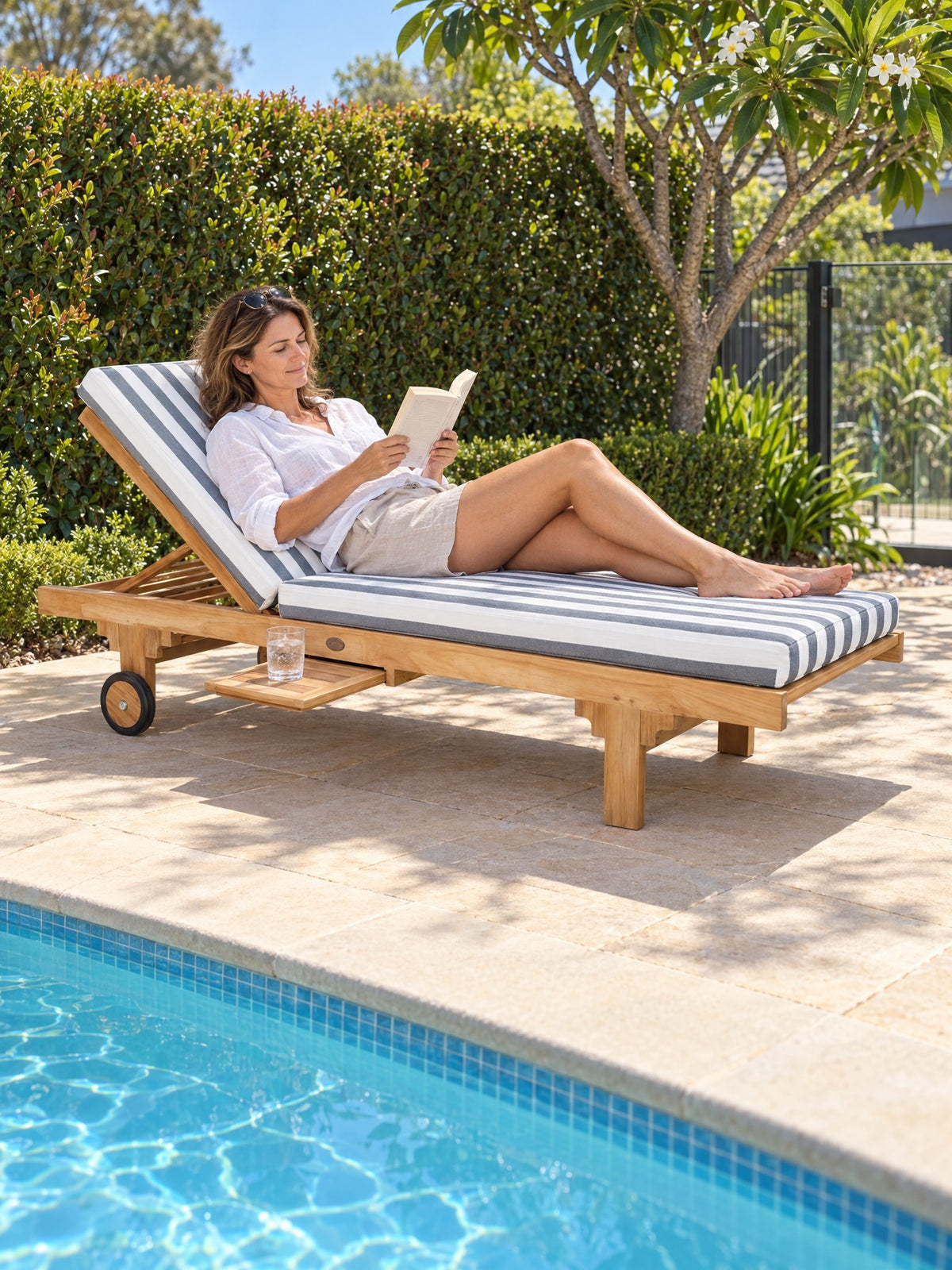 Woman reading on a Traditional teak pool lounge with a denim and white stripe cushion beside a pool