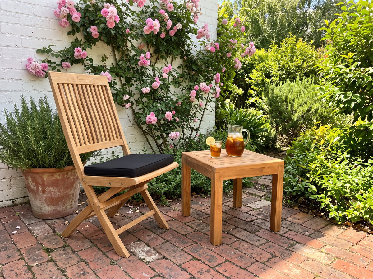 Teak Capri folding chair with black cushion and a square teak side table in a courtyard with climbing roses