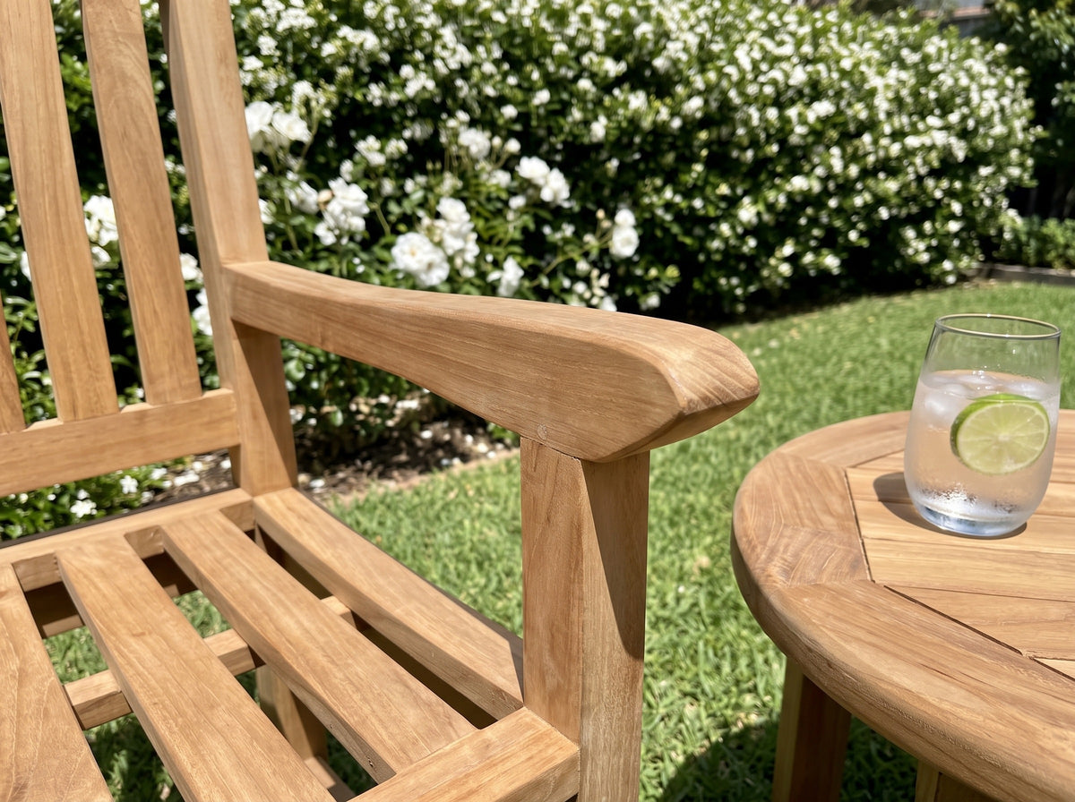 Close-up of the broad armrest of a teak Sierra armchair beside a round teak side table in a sunny garden