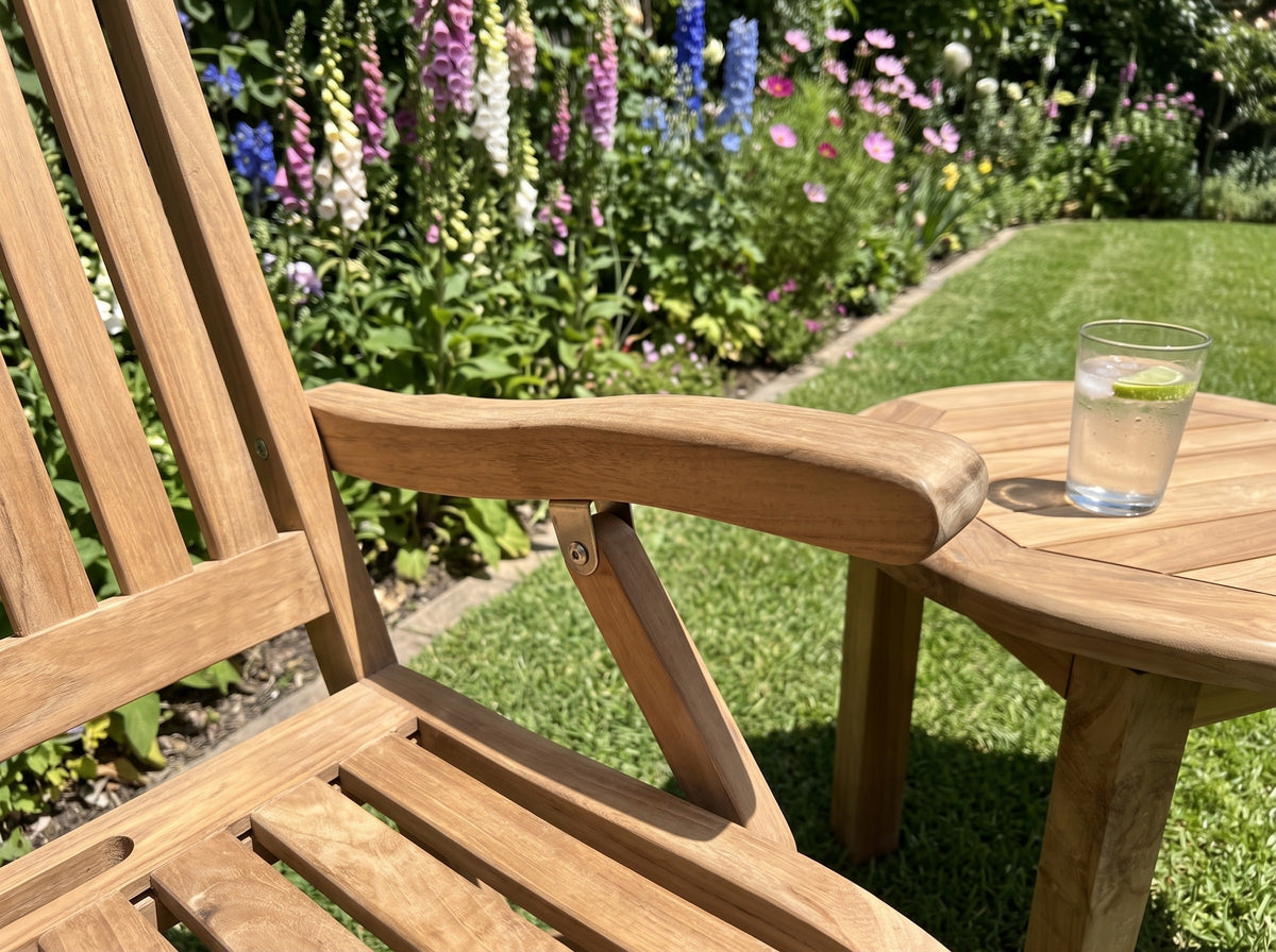 Close-up of the armrest of a teak Vivienne folding armchair beside a round teak side table in a cottage garden