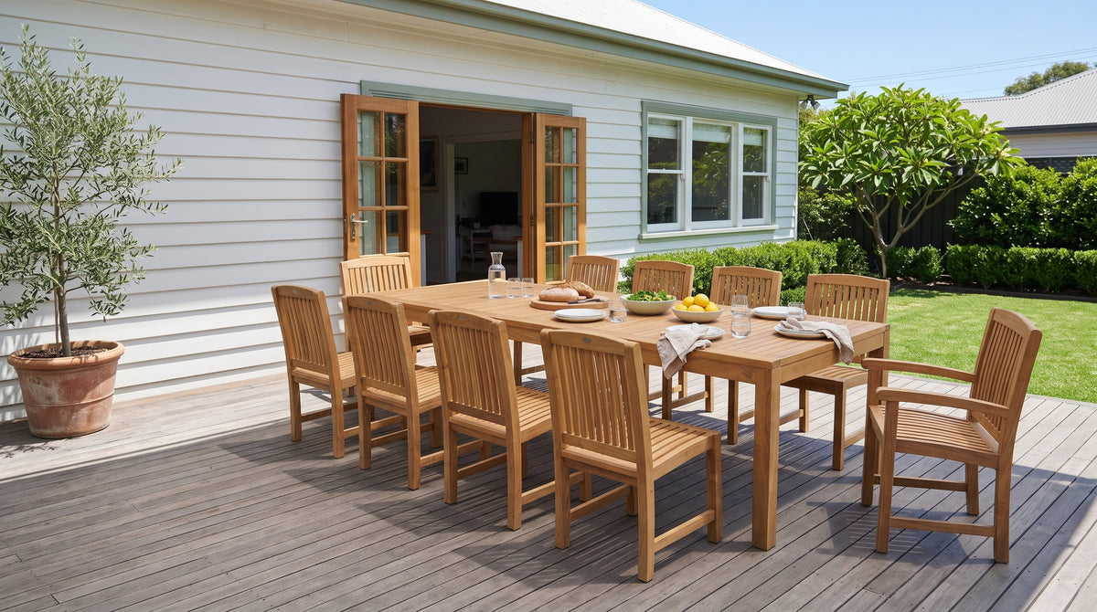 Teak dining table with ten Japan chairs on a timber deck behind a white weatherboard house, set for lunch in summer sun